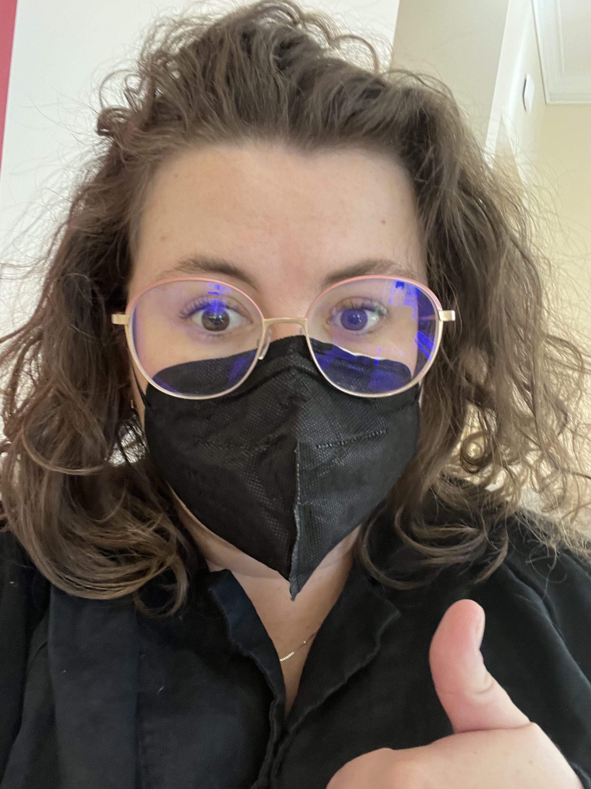 Emma wearing a black facemask, giving a thumbs up. She has glasses and is wearing a black collared shirt.