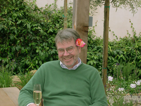 Edward English sitting at a table with a glass of champagne with greenery in the background and a rose tucked behind his ear
